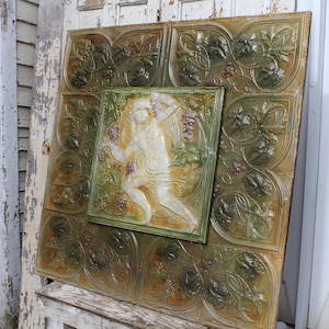 May include: A vintage tile panel with a green and brown floral design. A central tile features a white figure of a woman with flowing hair and a flowing dress. The tile is surrounded by a border of green and brown leaves.