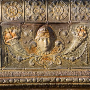 May include: Close-up of a weathered, ornate metal panel with a repeating pattern. The panel features embossed floral and geometric designs, along with a central face motif. The color palette is primarily shades of brown and gold, with areas of rust.