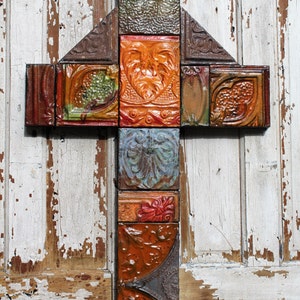 May include: A handcrafted cross composed of various rectangular tiles. The tiles feature embossed designs in shades of orange, green, brown, and blue. The cross is mounted on a distressed, white-painted wooden door.