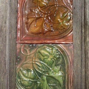 May include: Two square ceramic tiles with a floral design. The top tile is orange, brown, and green. The bottom tile is green, brown, and pink.