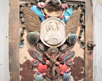 May include: A mixed media artwork featuring a central image of the Virgin Mary, surrounded by seashells, butterflies, and other decorative elements. The artwork is mounted on a wooden panel with a distressed finish.