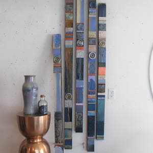 Peut inclure: Une installation murale d'art composée de six panneaux verticaux en bois, chacun composé de différentes tuiles bleues, grises et brunes. Les panneaux sont disposés en quinconce, créant une œuvre visuellement intéressante et texturée.