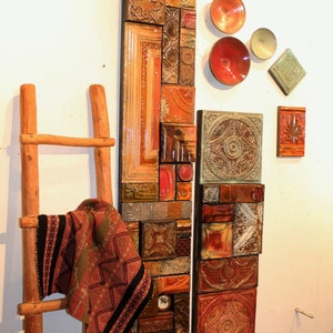 May include: A wall art piece made of various tiles in shades of brown, orange, and green. The tiles are arranged in a vertical pattern and are framed by a wooden frame. A wooden ladder with a red and black patterned blanket draped over it is in the foreground.