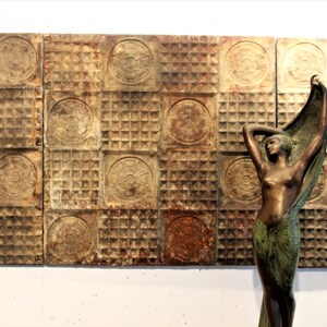 May include: A bronze-toned sculpture of a standing figure with arms raised, draped in a green cloth, positioned in front of a wall panel composed of weathered, square metal tiles featuring alternating circular floral motifs and grid patterns.