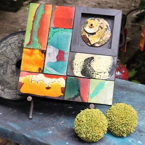 May include: A colorful abstract mosaic artwork made of ceramic tiles, framed in black wood, and displayed on a black easel. The artwork features a variety of colors, including red, orange, yellow, green, blue, and black. The tiles are arranged in a geometric pattern, and the artwork is accented with a round, yellow ceramic tile in the center.