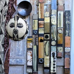 Puede incluir: Una composición artística que presenta una máscara tallada con patrones en blanco y negro, un cuenco de metal oscuro y una colección de baldosas de cerámica texturizadas en varios colores. Un textil estampado cuelga a la izquierda.