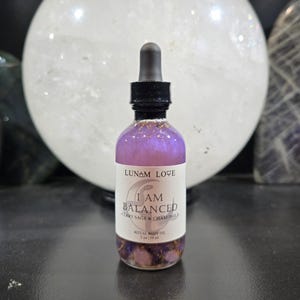 May include: A clear glass bottle with a black dropper top, filled with a purple liquid. The label reads "Lunam Love I Am Balanced Clary Sage & Chamomile Ritual Body Oil 2 oz / 59 ml."