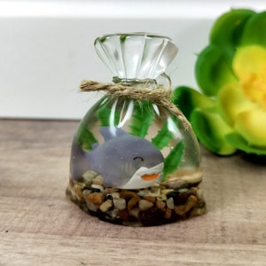 May include: A clear glass bag with a rope tie, filled with water, green plants, and a small gray shark figurine. The bag is sitting on a wooden surface.