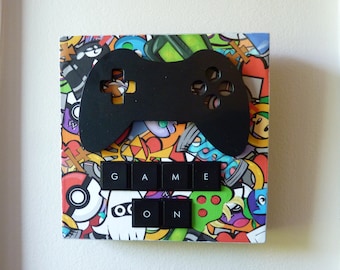 Peut inclure: Une décoration murale carrée présentant un collage coloré d'icônes de dessins animés et de jeux vidéo. Une découpe noire de manette de jeu est fixée par-dessus, avec six touches de clavier d'ordinateur noires formant les mots GAME ON en dessous.