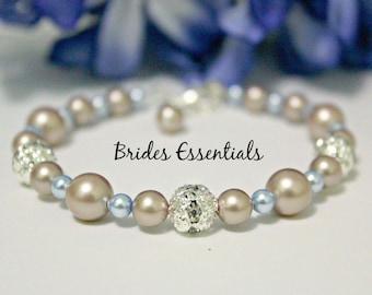 May include: A beaded bracelet featuring large, taupe-colored faux pearls interspersed with smaller, light blue beads and sparkling silver accents. The bracelet has the text "Brides Essentials" on it.