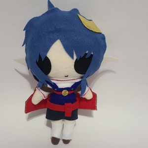 May include: A blue and white plush toy with a red cape and a gold crown. The toy has large black eyes and a small, smiling mouth.