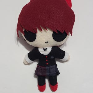 Puede incluir: Un juguete de peluche de un personaje con cabello rojo, ojos negros y un atuendo blanco y negro. El juguete lleva una falda de cuadros negros y rojos y zapatos rojos.