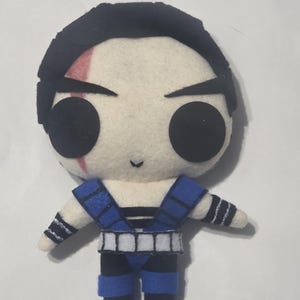 May include: A handmade felt doll of a character with a pale face, black hair, and large black eyes. The doll wears a blue and black outfit with white accents. A red scar is visible on the character's face. The doll is a stylized representation of a video game character.