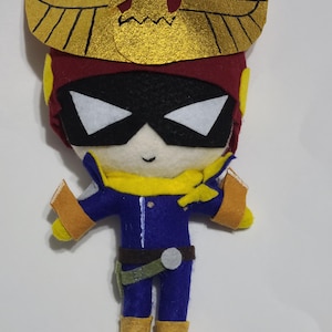May include: A felt plush toy of a character wearing a red and gold helmet with a winged emblem. The character is wearing a blue jumpsuit with a yellow scarf and gold boots.