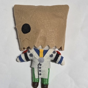 May include: A handmade felt doll with a brown paper bag head featuring a black circle and star cut-out. The doll wears a white coat with blue and black polka dots, green striped pants, and brown shoes. Red accents are on the shoulders.