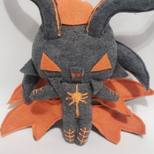 May include: A handmade plush doll in shades of gray and orange. The doll features large, pointed ears, triangular eyes, and a stylized sun design on its chest. The doll has a unique, whimsical design.