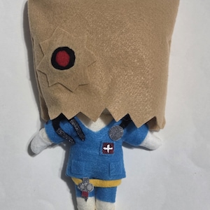 May include: A handmade felt doll with a brown paper bag head featuring a star-shaped cutout with a red and black center. The doll wears a blue felt medical uniform with a white cross emblem and a stethoscope.