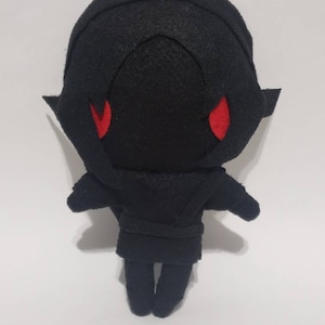 May include: Black plush toy with a hooded cloak and red eyes. The toy is standing on a white surface.