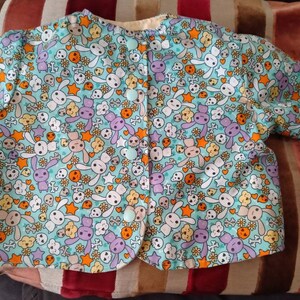 May include: A light blue baby cardigan with a button closure. The cardigan is covered in a whimsical print of bunnies, skulls, stars, and flowers.