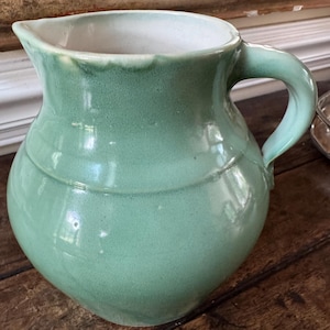 May include: A light green ceramic pitcher with a rounded body, a wide spout, and a curved handle. The exterior features a smooth, glossy glaze with subtle variations in color, while the interior is finished in a contrasting white glaze.