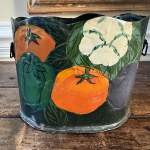 May include: A metal bucket with a dark green background and a wavy top edge. The bucket is painted with colorful vegetables, including orange tomatoes, a green bell pepper, and a white cauliflower. Black handles are on either side.