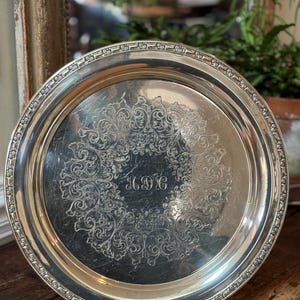May include: A round, silver-colored serving tray with an ornate floral design in the center and along the rim. The tray has a reflective surface and appears to be vintage. The initials "MDC" are engraved in the center of the tray.