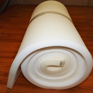 May include: A white foam roll, tightly rolled up, with a white tape around the center. The foam is used for upholstery and padding.