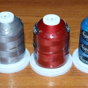May include: Ten spools of thread in various colors, including white, gray, red, blue, yellow, and brown. Each spool is labeled "Signature" and has a white plastic base.