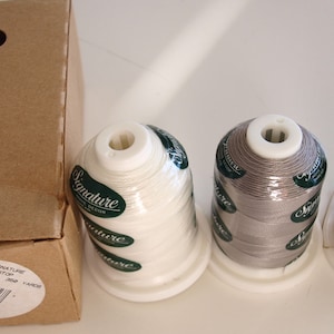 May include: Three spools of Signature thread in white, gray, and brown. The spools are in a cardboard box with a label that says "Signature Slunk Top 3000 Yards Poly".