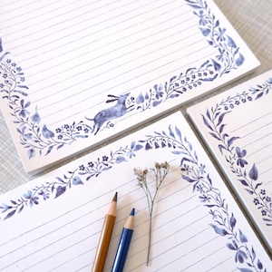 May include: White lined notepads with a blue floral border and a running hare illustration. The pads have a watercolour design with a blue and white colour scheme. Two pencils and a sprig of dried flowers are in the foreground.
