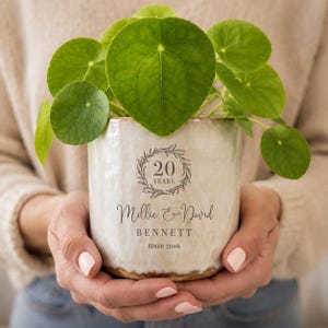 May include: A cylindrical ceramic planter with a cream-coloured, textured finish. The front features a printed design with a laurel wreath surrounding the text 20 YEARS, followed by the names Millie & David BENNETT and the text Since 2006.
