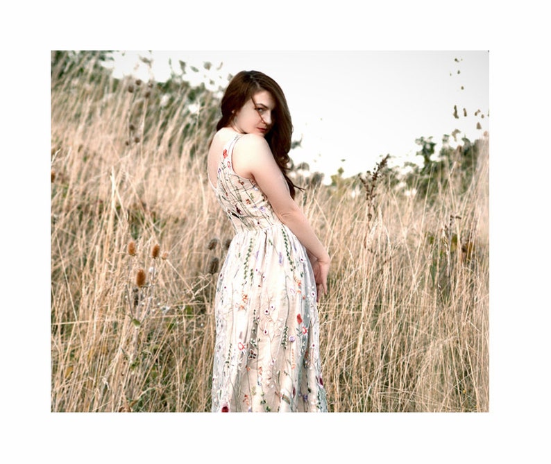 May include: A woman in a white dress with a floral pattern stands in a field of tall grass. She is looking over her shoulder and has her hair pulled back.