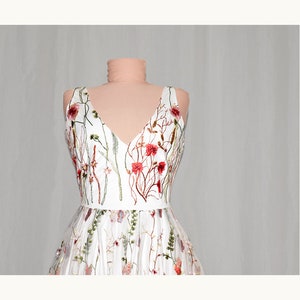 May include: A white sleeveless dress with a V-neckline and floral embroidery. The embroidery features red, pink, and green flowers and leaves.