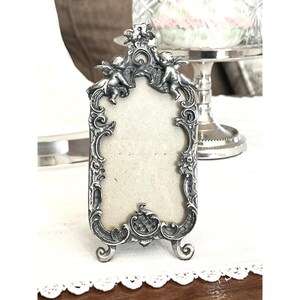 May include: A silver-toned metal picture frame with an ornate, rectangular design featuring two cherubs at the top. The frame has intricate scrollwork and floral detailing along the edges, supported by two small, curved feet at the base.