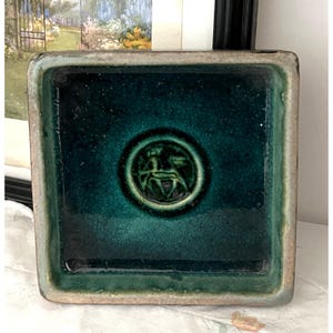 May include: A square ceramic dish featuring a deep teal, crackled glaze finish. The center of the dish displays a circular medallion with a raised relief design of a figure holding a bow and arrow, surrounded by a lighter green border.
