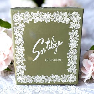 May include: A green box with white floral lace trim and the word "Sortilège" in elegant script. The text "LE GALION" is below the script. The box is surrounded by pink flowers.