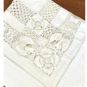 May include: A white, rectangular tablecloth with intricate lace detailing. The lace features floral and geometric patterns, creating a delicate and elegant design. The tablecloth is made of a soft, woven fabric.