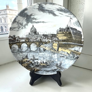 May include: A round ceramic decorative plate featuring a detailed black and white ink-style illustration of a cityscape with a large domed building, a stone bridge over a river, and a fortress, accented with muted yellow and blue tones.