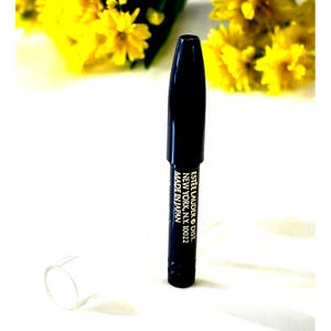 May include: A black eyeliner pencil with a clear cap, with text that reads "ESTEE LAUDER DIST. NEW YORK, N.Y. 10022 MADE IN JAPAN". The background features yellow flowers.