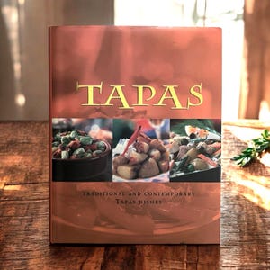 May include: A cookbook titled "TAPAS" with a reddish-brown cover. The title is in gold lettering. Below the title are three images of tapas dishes. The text "TRADITIONAL AND CONTEMPORARY TAPAS DISHES" is below the images.
