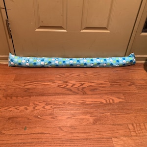 May include: A long, cylindrical door draft stopper made of fabric. The stopper is light blue with a repeating pattern of white, green, and blue floral and circular designs. It rests on a wooden floor in front of a closed door.