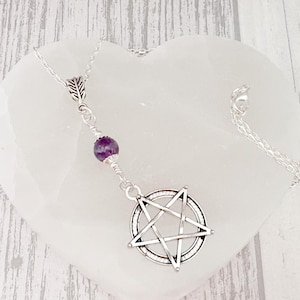 May include: A silver-toned pendant necklace featuring a pentagram charm and a small purple gemstone. The necklace is displayed on a white heart-shaped dish, set against a light wood-grain background. The pendant is a symbol of Wicca and Paganism.