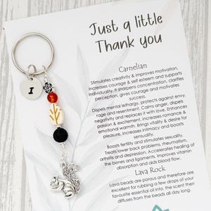 May include: A silver keyring with a squirrel charm, colourful beads, and the letter "I". The card reads "Just a little Thank you" and details the properties of Carnelian and Lava Rock.