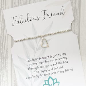 May include: A silver beaded bracelet with a heart charm is presented on a white card. The card is inscribed with "Fabulous Friend" and a message about friendship. A logo and website address are also visible on the card.