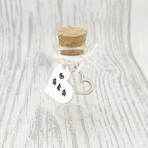 May include: A small, clear glass bottle with a cork stopper. A white heart-shaped tag with the word "mum" and heart symbols hangs from the bottle. A silver heart charm is also attached. The bottle is a decorative item.