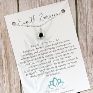 May include: A silver-toned wire-wrapped necklace featuring a small black teardrop-shaped stone. The necklace is displayed on a white card labeled Empath Barrier, Black Tourmaline, and Meaningful Charm with descriptive text and a lotus flower logo.