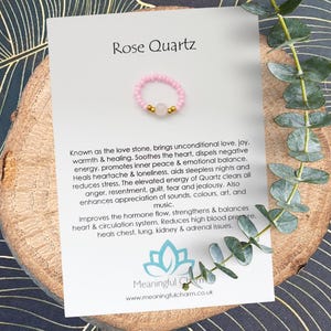 May include: A pink Rose Quartz ring with a small, round, light pink stone and gold-colored beads. The ring is displayed on a wooden surface with a card that reads "Rose Quartz" and details about the stone's properties.