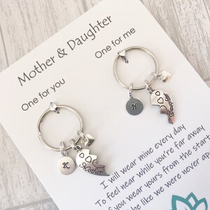 May include: Two silver keychains with heart charms and a broken heart charm that says "Mother" and "Daughter". The keychains are on a white card with the text "Mother & Daughter" and a poem about wearing the keychains to feel close to each other.
