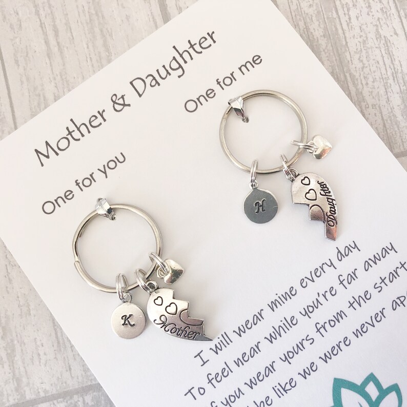 May include: Two silver keychains with heart charms and a broken heart charm that says "Mother" and "Daughter". The keychains are on a white card with the text "Mother & Daughter" and a poem about wearing the keychains to feel close to each other.