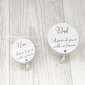 May include: Two white, round memorial ornaments with silver rims and white string. One says "Nan" and the other says "Dad", both with the text "A piece of you is with me forever" and a heart symbol. Each has a small feather charm.